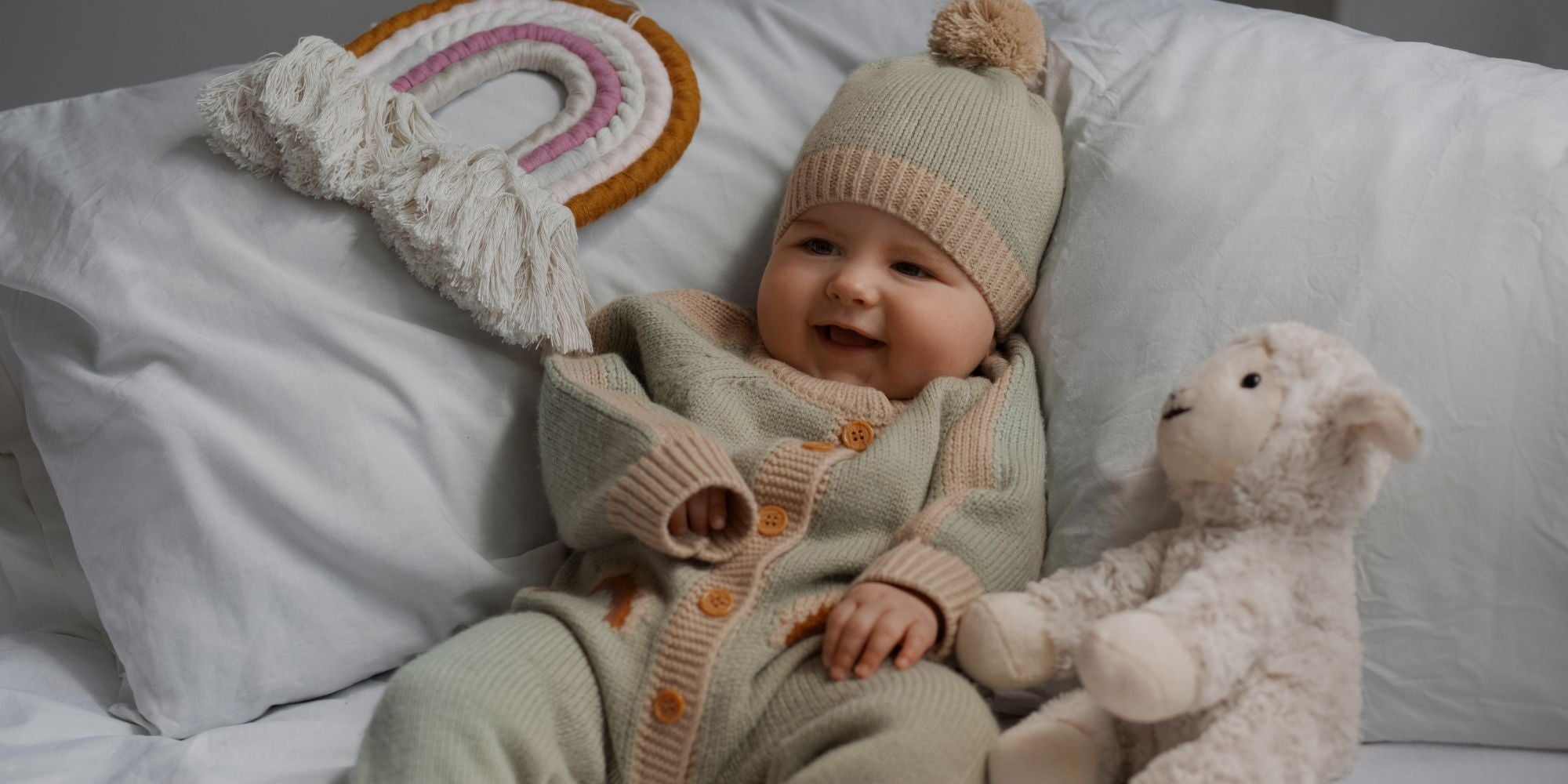 A baby wearing a cozy knit beige and sage green outfit with a matching beanie and pom-pom, lying on white pillows next to a plush sheep toy and a crocheted rainbow.